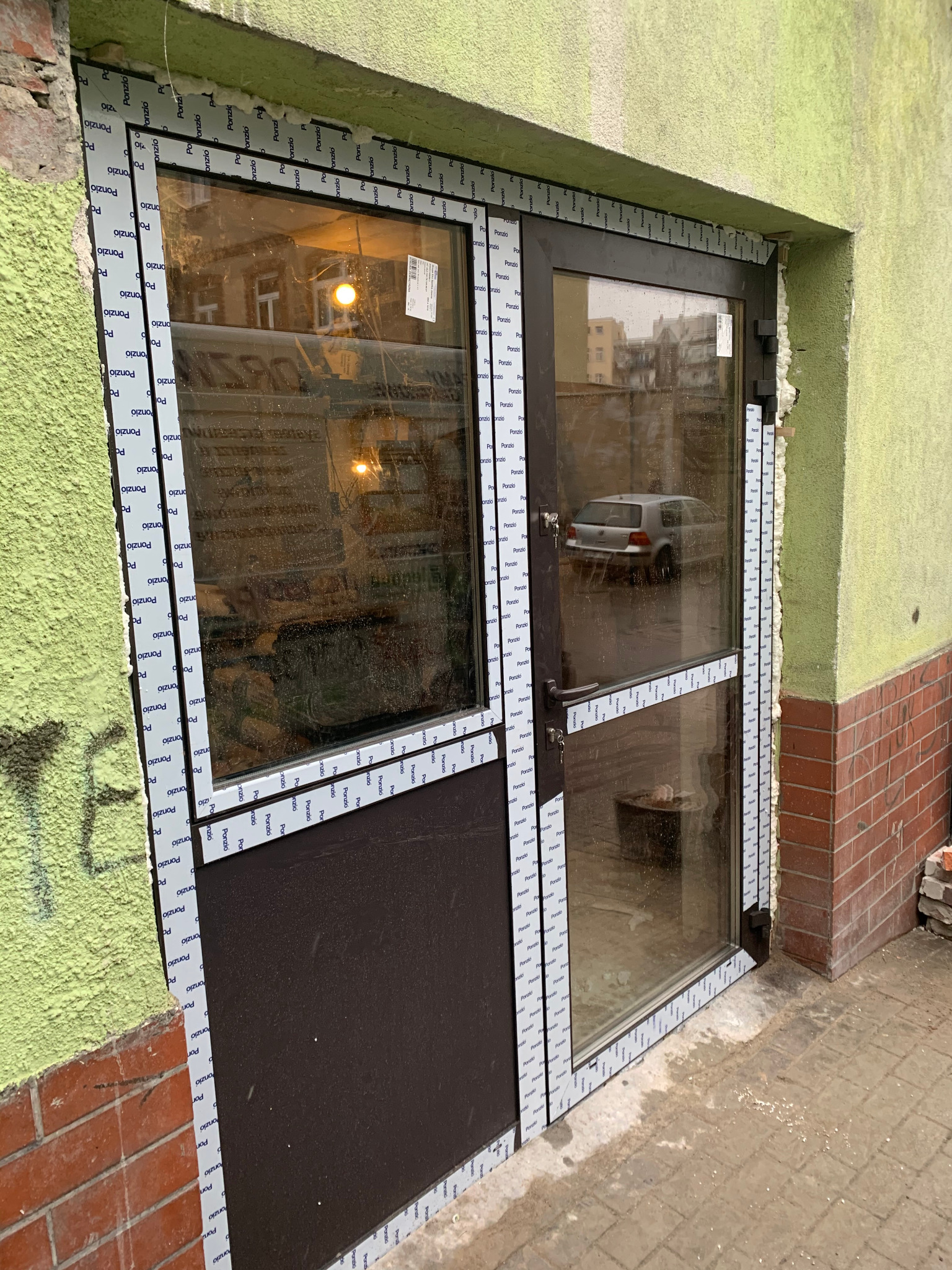 Montaż dwuskrzydłowych drzwi z aluminiową ramą, widoczne pianowanie i taśmy zabezpieczające. Częściowo przeszklone, z brązowym panelem dolnym. Budynek w tle.