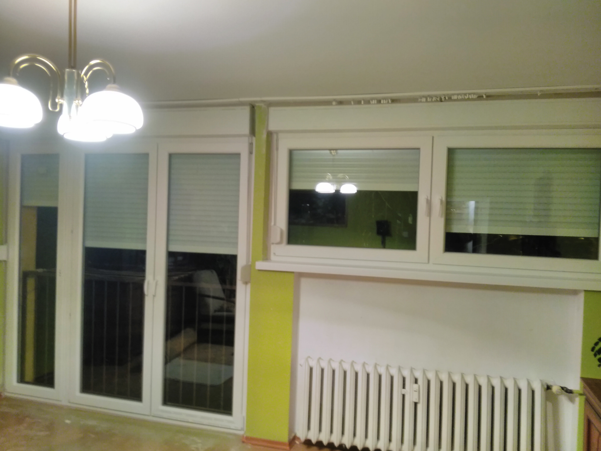 Białe okna z plisami w pokoju z grzejnikiem i lampą sufitową. Widoczne trzy okna: jedno potrójne balkonowe i dwa mniejsze, pojedyncze. Ściany w odcieniach bieli i limonki.