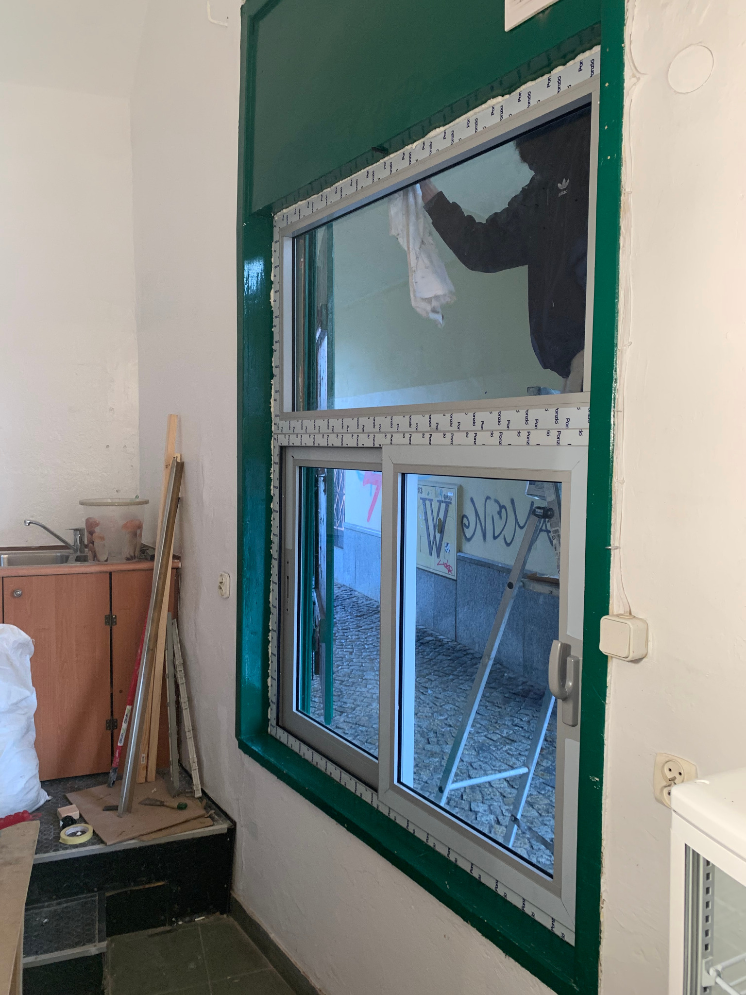 Montaż okna PCV z zieloną ramą. Osoba czyści szybę od wewnątrz. Widoczne narzędzia i drabina. Ujęcie z boku, wnętrze pomieszczenia w trakcie prac wykończeniowych.