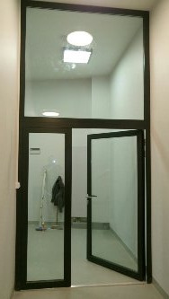 Szklane drzwi z czarną ramą, otwarte do wewnątrz, z górnym naświetlem, w jasnym korytarzu z widocznym oświetleniem sufitowym i elementami garderoby w tle.