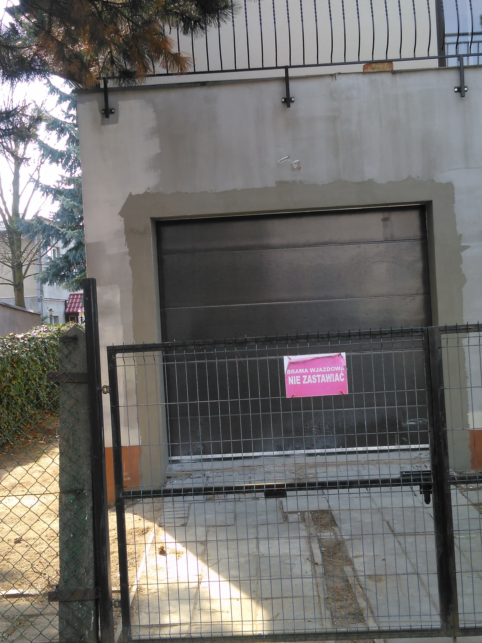 Brama garażowa segmentowa w surowym otoczeniu, widoczna przez metalowe ogrodzenie z różową kartką 'Brama wjazdowa - nie zastawiać'.
