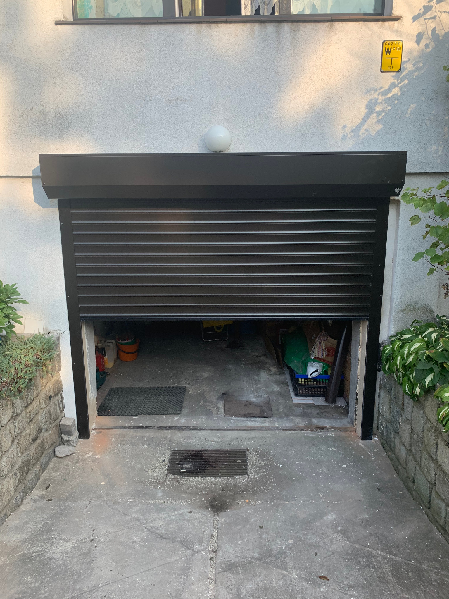 Częściowo otwarta, czarna brama rolowana garażowa z poziomymi panelami, widoczna zawartość garażu, ściana z tabliczką informacyjną i roślinność po bokach.
