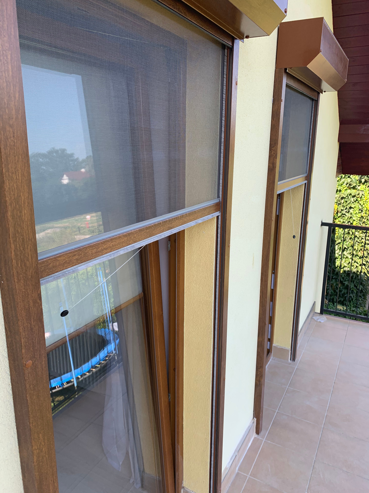 Dwa okna z brązowymi ramami i moskitierami na balkonie, widok z zewnątrz. Na szybach odbija się niebieski basen. Żółta elewacja budynku.