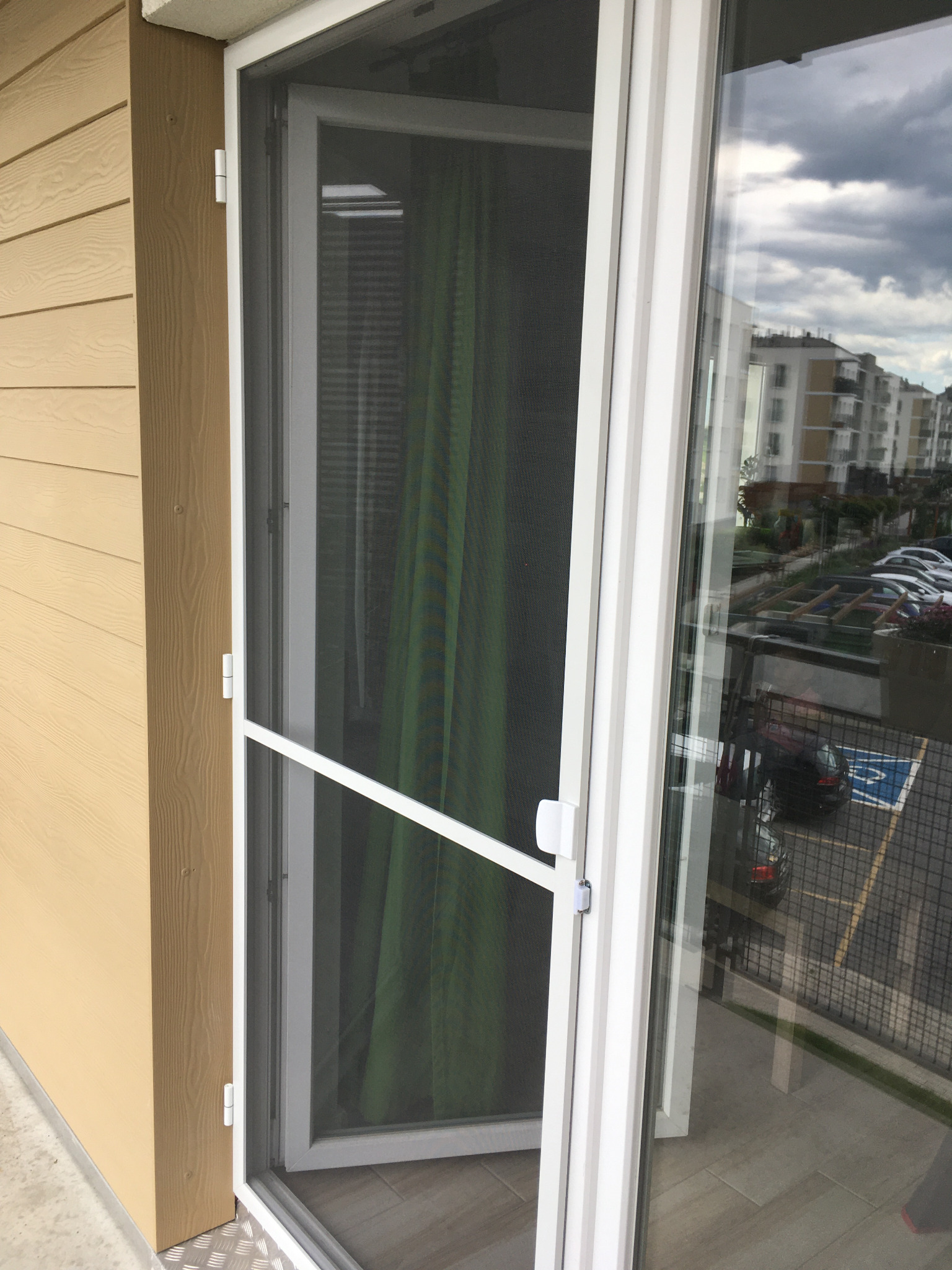 Biała rama okienna z moskitierą na tle budynku mieszkalnego i parkingu. Widoczne detale mocowania moskitiery i odbicie otoczenia w szybie.