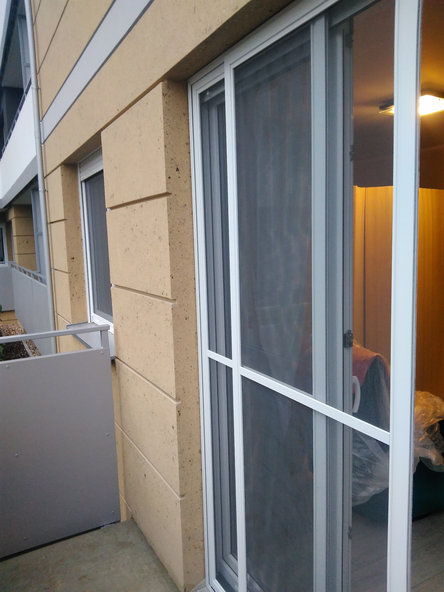 Białe, aluminiowe drzwi przesuwne z siatką moskitierą, widoczne z zewnątrz balkonu. W tle fragment elewacji budynku z oknem.