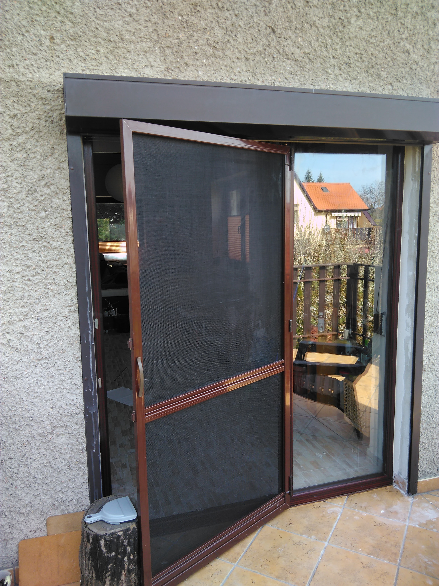 Otwarta moskitiera w brązowej ramie, zamontowana na balkonie z widokiem na dom z czerwoną dachówką. Na pierwszym planie pień z narzędziem.