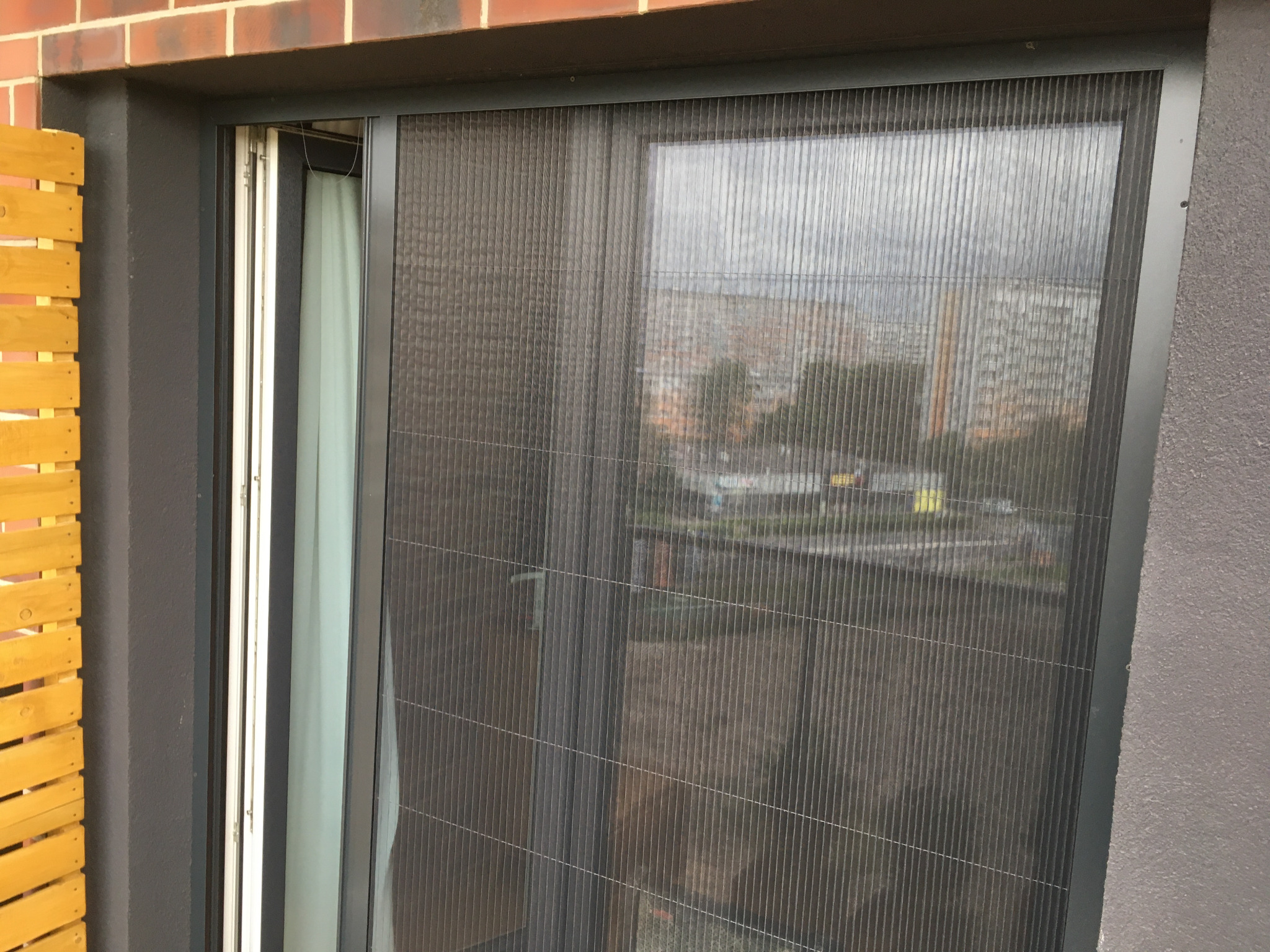Zamknięta, szara moskitiera plisowana na oknie balkonowym, z widokiem na budynki mieszkalne w tle. Widoczny fragment drewnianego ogrodzenia balkonu.