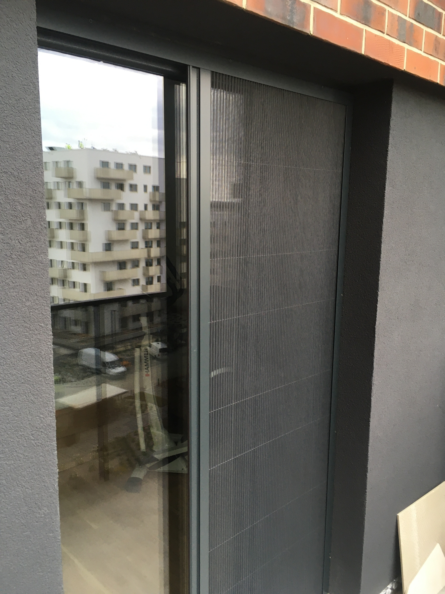 Szara plisa okienna zamontowana w oknie balkonowym, widok z bliska. W tle budynek mieszkalny odbijający się w szybie. Minimalistyczny design, nowoczesne wykończenie.