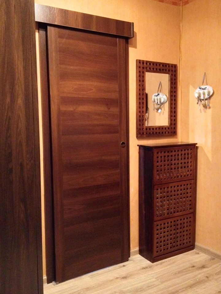 Ciemnobrązowe drzwi przesuwne z drewnianym wykończeniem, dopasowane do szafki na buty i ramy lustra w przedpokoju o ciepłej, pomarańczowej kolorystyce ścian.