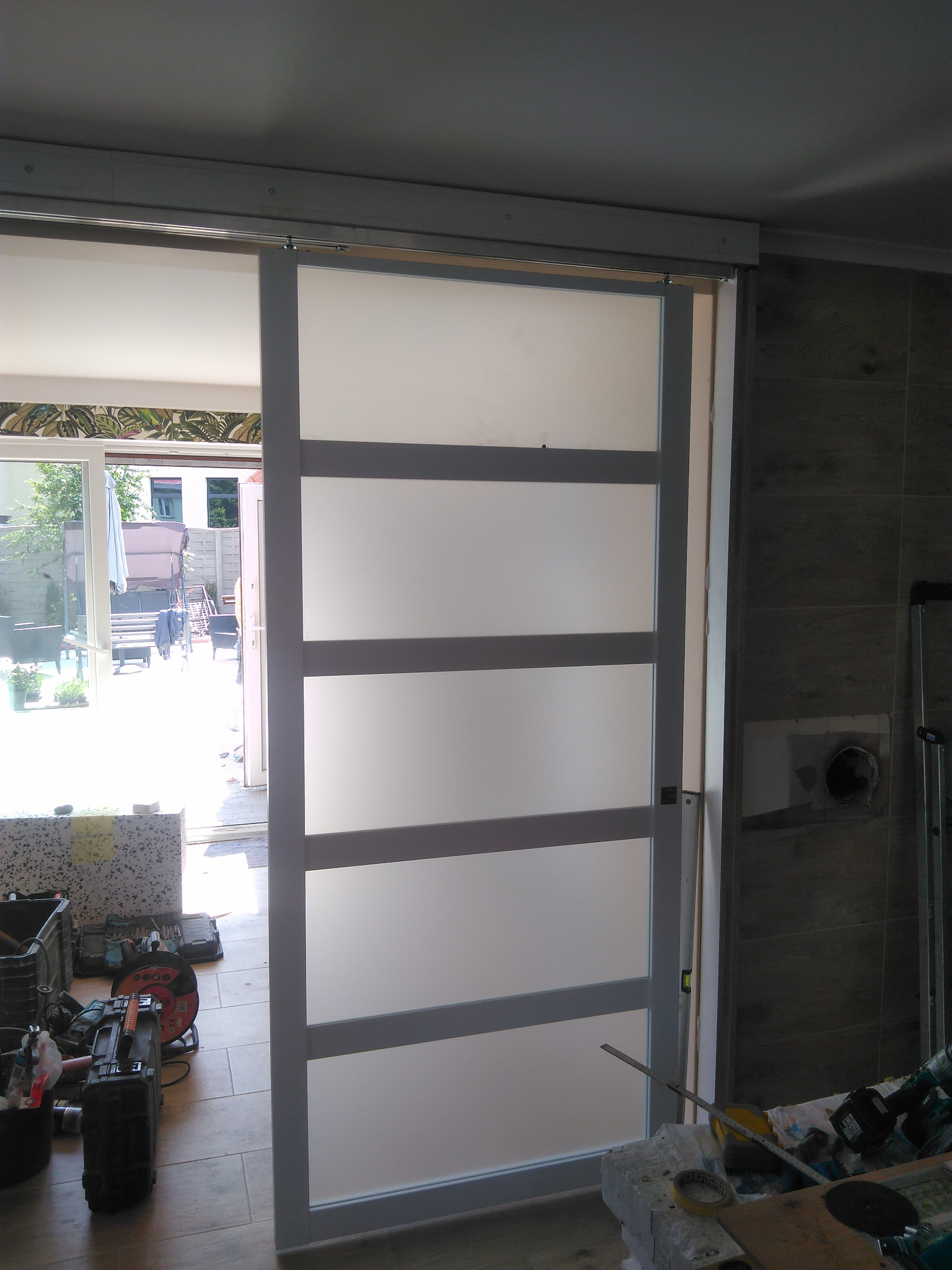 Zamontowane drzwi przesuwne z czterema matowymi panelami, widoczne narzędzia i fragment wnętrza z wyjściem na zewnątrz. Ujęcie z bliska, z widocznymi detalami montażu.