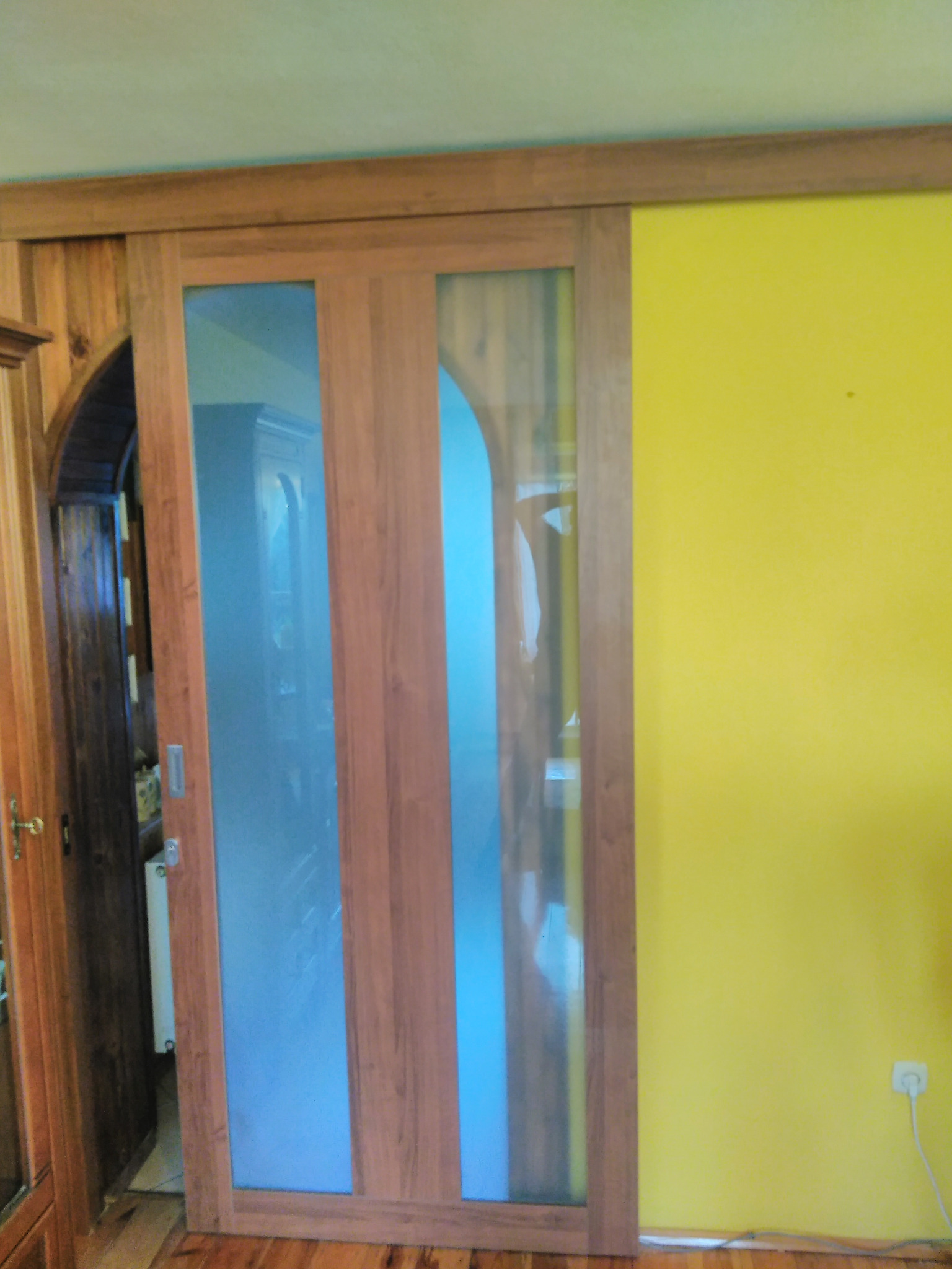 Drewniane drzwi przesuwne z dwoma szklanymi panelami, widoczne na tle żółtej ściany i fragmentu drewnianej boazerii. Widoczny fragment wnętrza za drzwiami.