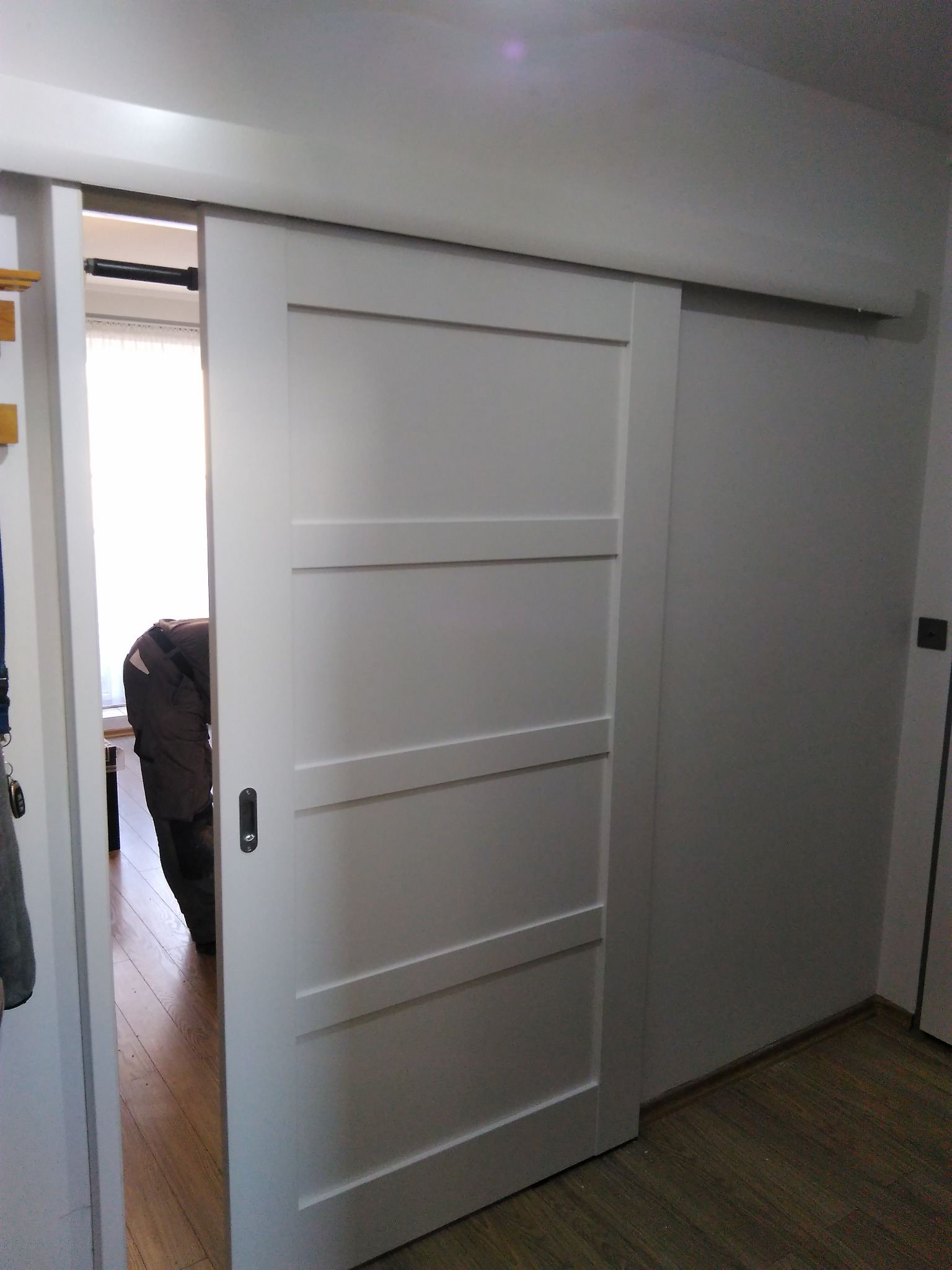 Białe, przesuwne drzwi z czterema panelami, częściowo otwarte, ukazujące fragment wnętrza z drewnianą podłogą i osobą w środku. Minimalistyczny design, jasne oświetlenie.