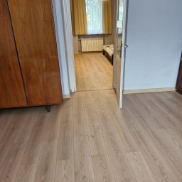 KK Studiokoloru - Jasne panele podłogowe o wzorze drewna w pokoju z błękitną ścianą i starym drewnianym kredensem, widok przez otwarte drzwi na sypialnię.