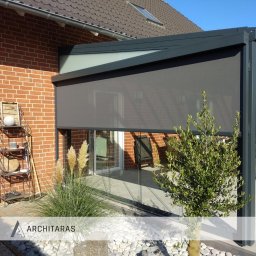 Architaras D.Paprocki Sp. z.o.o. - Zadaszenie tarasu z szarą roletą, ceglana ściana budynku, dekoracyjna półka z ozdobami i trawy ozdobne. Nowoczesna konstrukcja z elementami szklanymi.