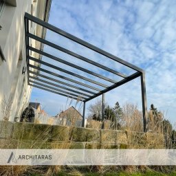 Architaras D.Paprocki Sp. z.o.o. - Nowoczesna, grafitowa konstrukcja zadaszenia tarasu z widokiem na błękitne niebo i zabudowę miejską w tle. Zadaszenie wsparte na kamiennym murku.