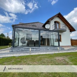 Architaras D.Paprocki Sp. z.o.o. - Szklana konstrukcja z ciemną ramą, dobudowana do domu z balkonem. Widoczne schody, trawnik i dekoracyjne oświetlenie zewnętrzne. Wnętrze z meblami.