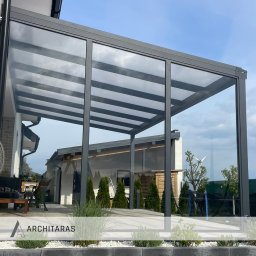 Architaras D.Paprocki Sp. z.o.o. - Nowoczesna, szara konstrukcja z przezroczystym zadaszeniem, przylegająca do budynku. Widoczne meble ogrodowe i zadbana zieleń. Minimalistyczny design.