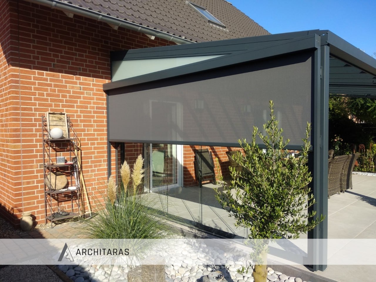 Zadaszenie tarasu z szarą roletą, ceglana ściana budynku, dekoracyjna półka z ozdobami i trawy ozdobne. Nowoczesna konstrukcja z elementami szklanymi.