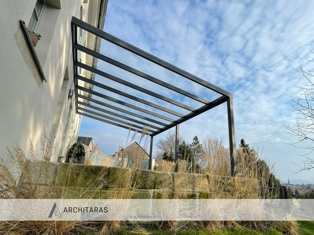 Nowoczesna, grafitowa konstrukcja zadaszenia tarasu z widokiem na błękitne niebo i zabudowę miejską w tle. Zadaszenie wsparte na kamiennym murku.