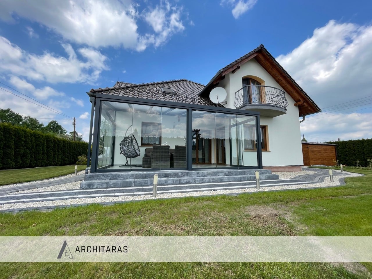 Szklana konstrukcja z ciemną ramą, dobudowana do domu z balkonem. Widoczne schody, trawnik i dekoracyjne oświetlenie zewnętrzne. Wnętrze z meblami.