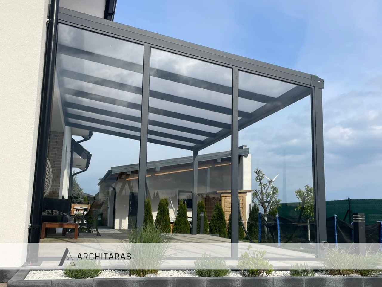 Nowoczesna, szara konstrukcja z przezroczystym zadaszeniem, przylegająca do budynku. Widoczne meble ogrodowe i zadbana zieleń. Minimalistyczny design.