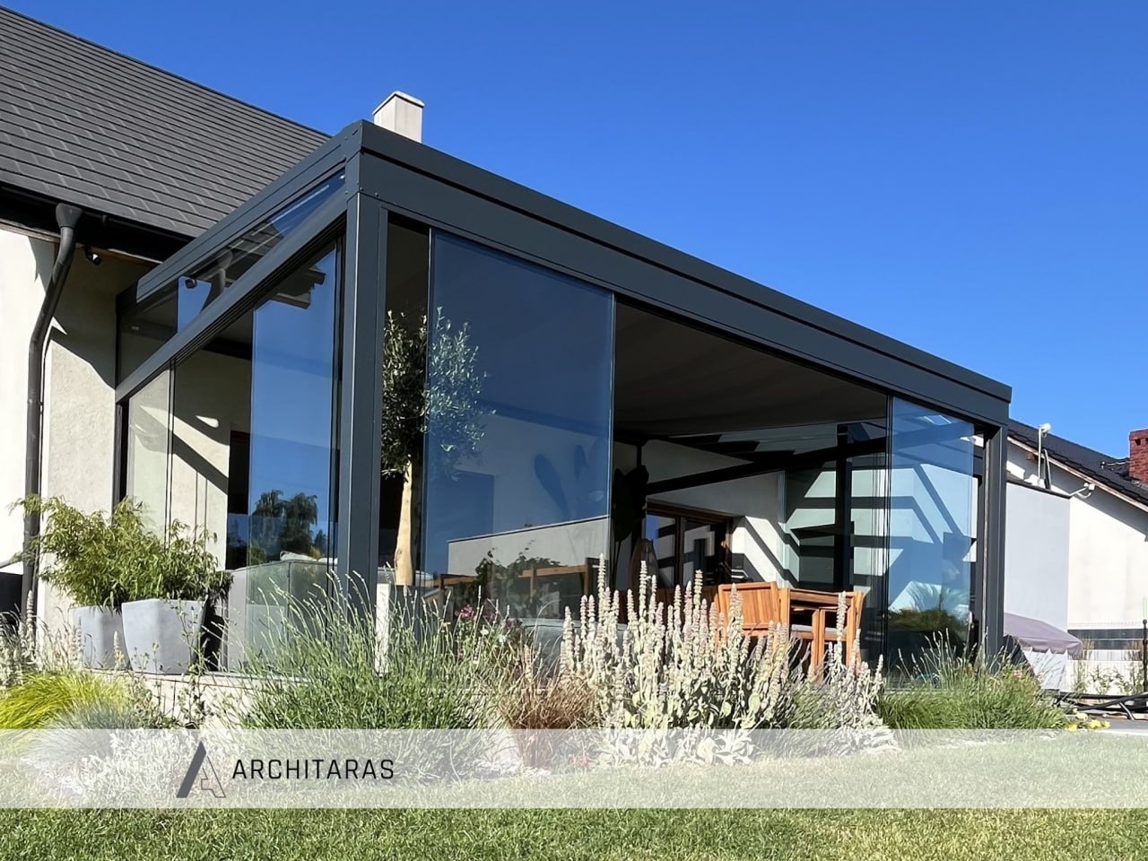 Nowoczesna, szklana konstrukcja z ciemną ramą, przylegająca do domu. Widoczne rośliny w donicach i elementy wyposażenia wnętrza. Błękitne niebo w tle.