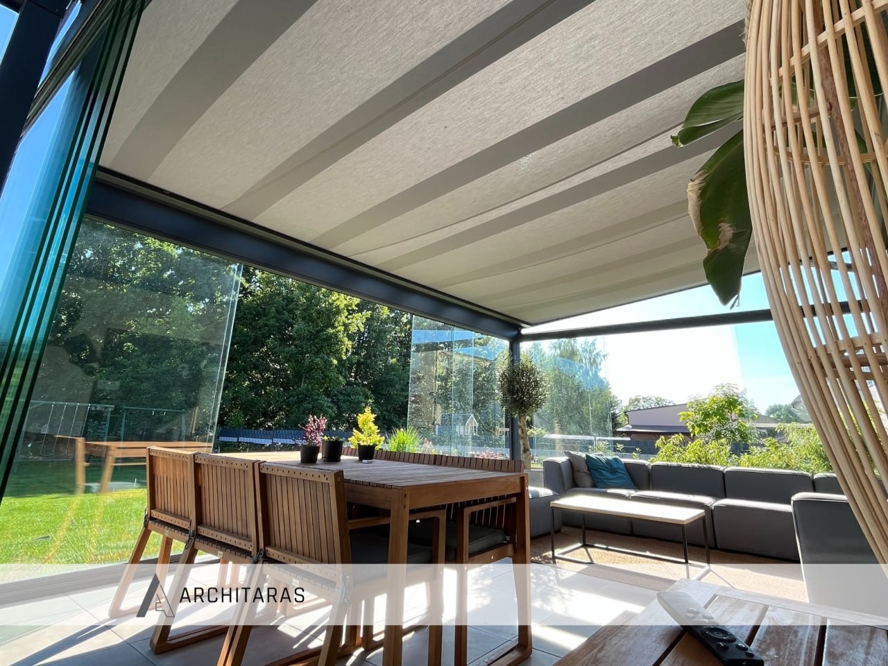 Zadaszony taras z przesuwnymi szklanymi ścianami, drewnianym stołem i szarą sofą. Widok na zielony ogród. Zadaszenie z regulowanymi panelami.