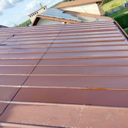 ŁUKASZ KOKOSZKA - Ceglasty dach z blachy trapezowej po renowacji, widok z góry. Na dachu panele solarne. Widoczne ślady wcześniejszych zabrudzeń na dolnej krawędzi.