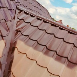 ŁUKASZ KOKOSZKA - Błyszczący, brązowy dach z blachodachówki modułowej, świeżo po renowacji, na tle błękitnego nieba z chmurami. Widoczne mocowania i detale wykończenia.