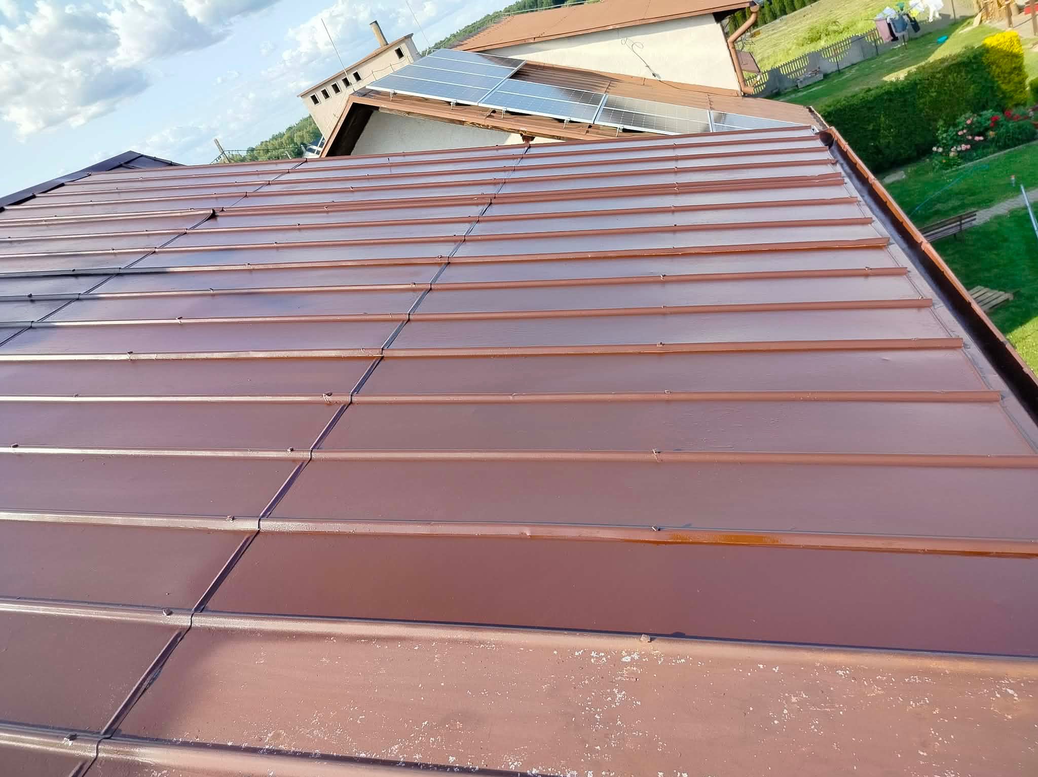 Ceglasty dach z blachy trapezowej po renowacji, widok z góry. Na dachu panele solarne. Widoczne ślady wcześniejszych zabrudzeń na dolnej krawędzi.