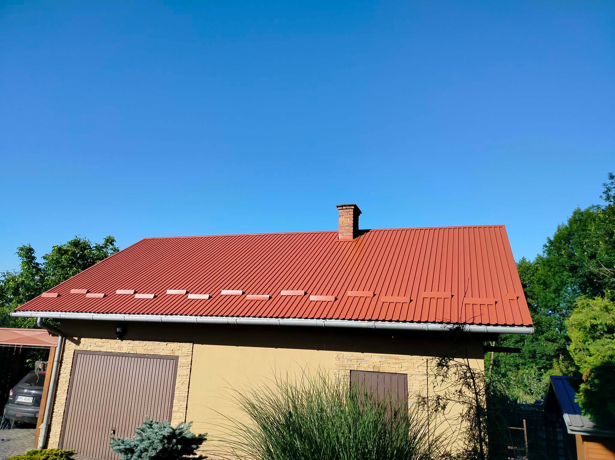 Czerwony, blaszany dach domu jednorodzinnego z ceglanym kominem na tle błękitnego nieba i zieleni drzew. Widoczny fragment elewacji z garażem.