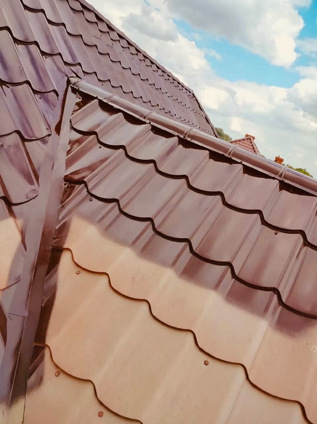 Błyszczący, brązowy dach z blachodachówki modułowej, świeżo po renowacji, na tle błękitnego nieba z chmurami. Widoczne mocowania i detale wykończenia.