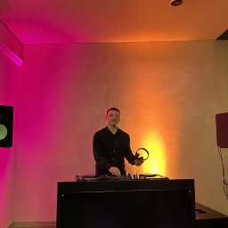 Mikolaj Mysliwiec - Uśmiechnięty DJ w czarnej koszuli, za konsoletą, trzyma słuchawki. Scena oświetlona kolorowymi światłami, tworząc klimat imprezy.