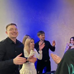 Mikolaj Mysliwiec - DJ z uśmiechem gra muzykę na imprezie, otoczony przez tańczących ludzi. Mikser Pioneer na pierwszym planie, niebieskie oświetlenie w tle.