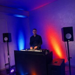 Mikolaj Mysliwiec - DJ za konsoletą, oświetlenie sceniczne w kolorach niebieskim i czerwonym, głośniki na statywach. DJ w czarnej koszuli, uśmiechnięty, w okularach.