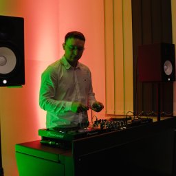 MIKO Y - DJ w okularach, w białej koszuli, obsługuje zielony mikser Pioneer w oświetlonym na czerwono i zielono pomieszczeniu. Widoczne głośniki studyjne.