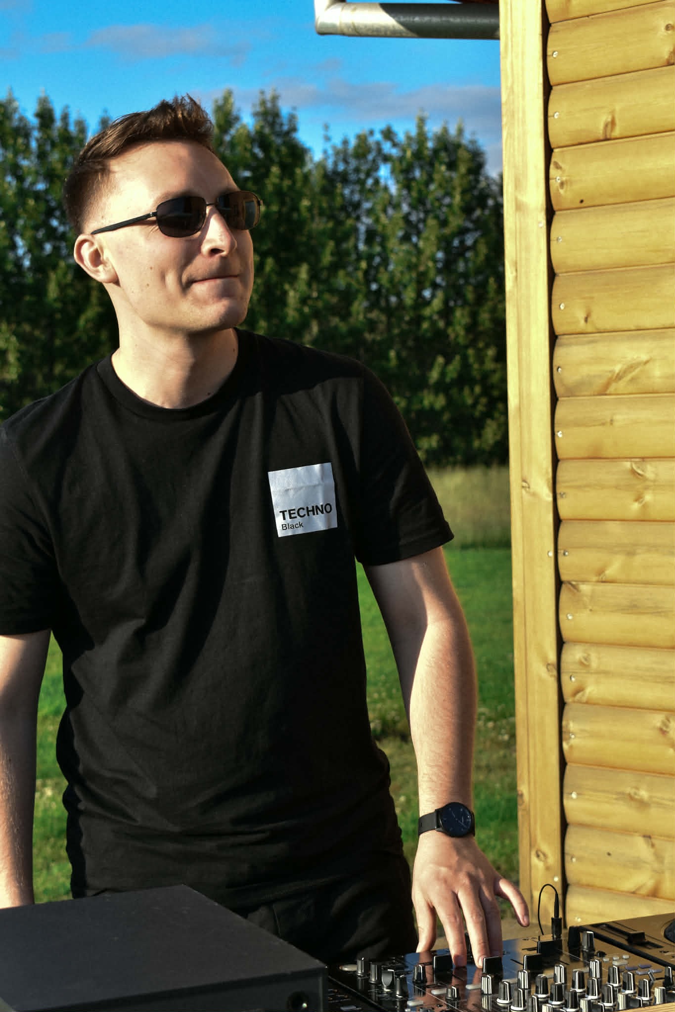 DJ w czarnej koszulce Techno Black, obsługujący mikser na tle drewnianej konstrukcji i zieleni. Ujęcie z boku, w okularach przeciwsłonecznych, w plenerze.