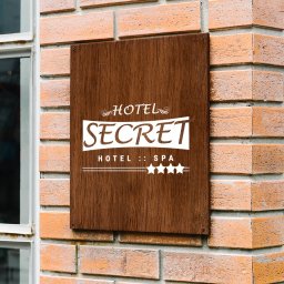 MaTaWi Marek Wojakowski - Drewniana tabliczka z logo 'Hotel Secret Hotel :: Spa' na ceglanej ścianie obok okna. Minimalistyczny design, ciepłe oświetlenie, wyraźna typografia.