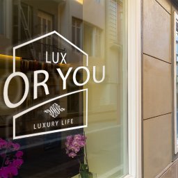 MaTaWi Marek Wojakowski - Naklejka z logo 'Lux For You Luxury Life' na witrynie sklepu, z odbiciem ulicy i kwiatami w tle. Minimalistyczny design w miejskim otoczeniu.