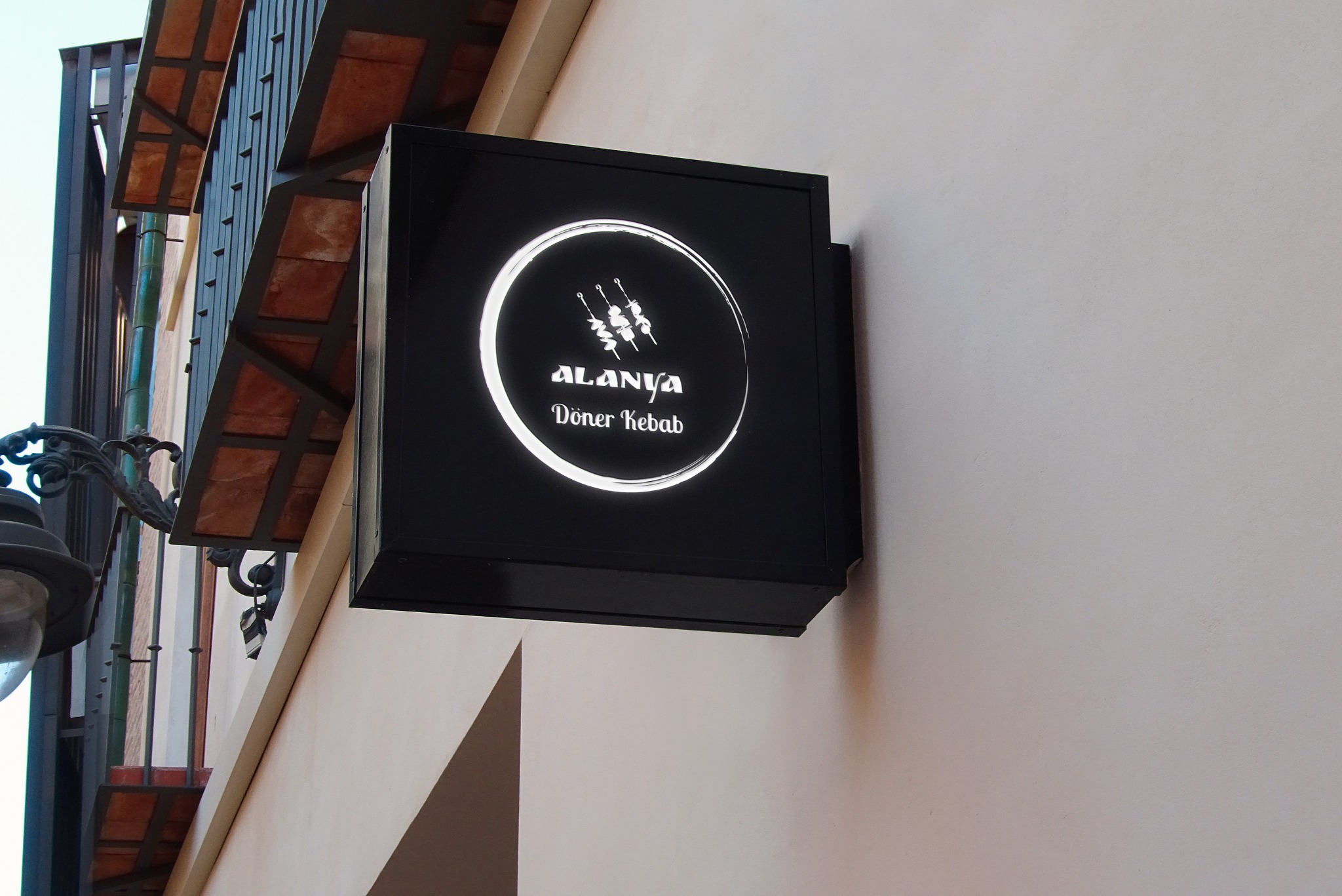 Kwadratowy, podświetlany szyld reklamowy 'Alanya Doner Kebab' na elewacji budynku, z widocznym fragmentem balkonu i lampy ulicznej w tle. Minimalistyczny design.