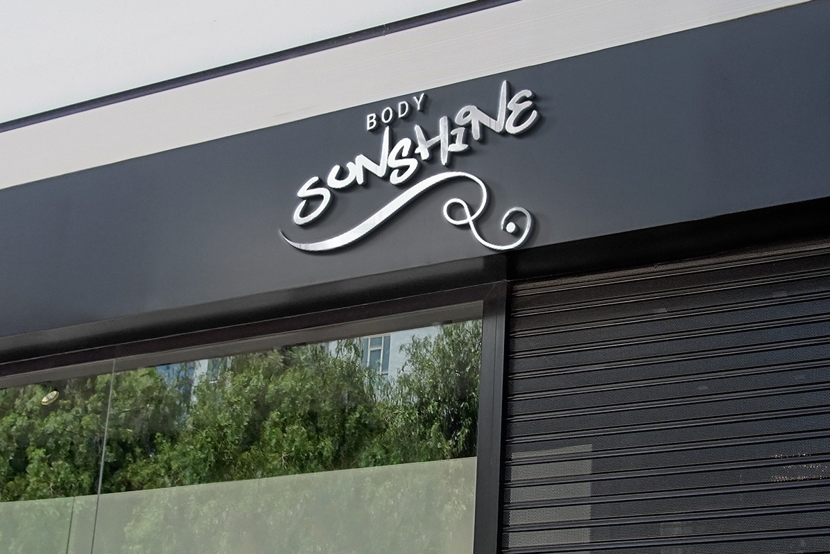 Srebrny, przestrzenny napis 'BODY SUNSHINE' nad witryną sklepu z opuszczoną roletą. W tle zieleń drzew odbijająca się w szybie.