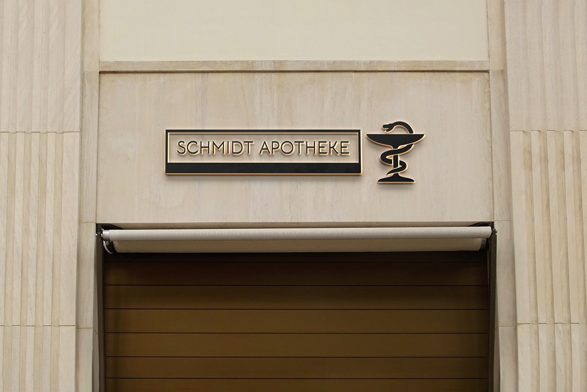 Elegancki szyld apteki 'Schmidt Apotheke' z symbolem Eskulapa, umieszczony nad wejściem z brązowymi drzwiami i kremową fasadą w stylu art deco.