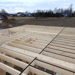 Follow River Sp. z o.o. - Drewniany szkielet podłogi domu w trakcie budowy, z widocznymi belkami i czarną izolacją, na tle krajobrazu z domami i drzewami w pochmurny dzień.