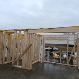 Follow River Sp. z o.o. - Wnętrze domu szkieletowego w trakcie budowy. Widoczne drewniane ramy ścian, folia izolacyjna, fragment podłogi i samochód w tle. Pochmurne niebo.