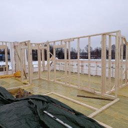 Follow River Sp. z o.o. - Wnętrze w trakcie budowy: drewniany szkielet domu z widoczną izolacją Eurovent, poziomica na folii ochronnej, zimowy krajobraz w tle. Etap wznoszenia ścian.