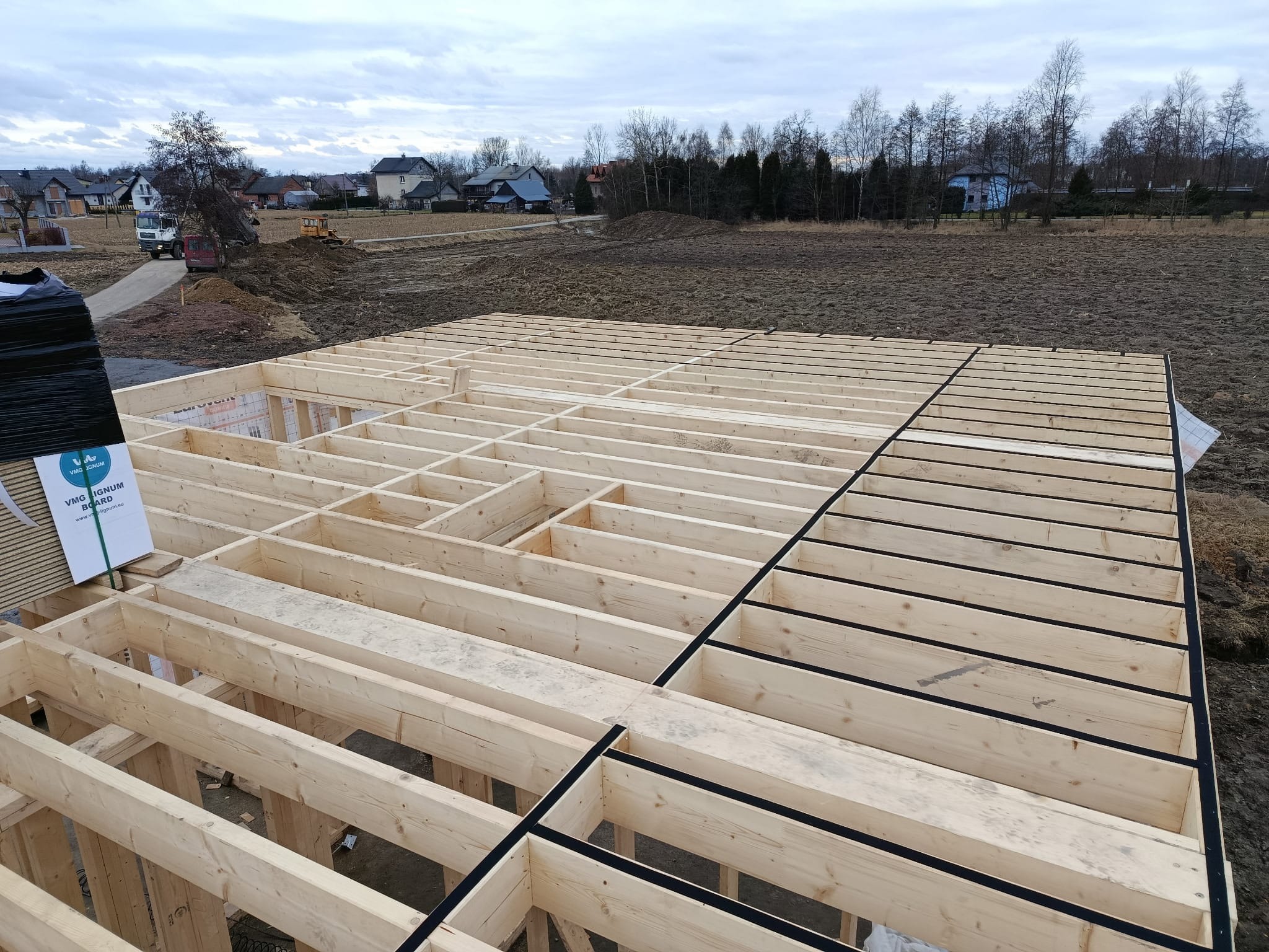 Drewniany szkielet podłogi domu w trakcie budowy, z widocznymi belkami i czarną izolacją, na tle krajobrazu z domami i drzewami w pochmurny dzień.