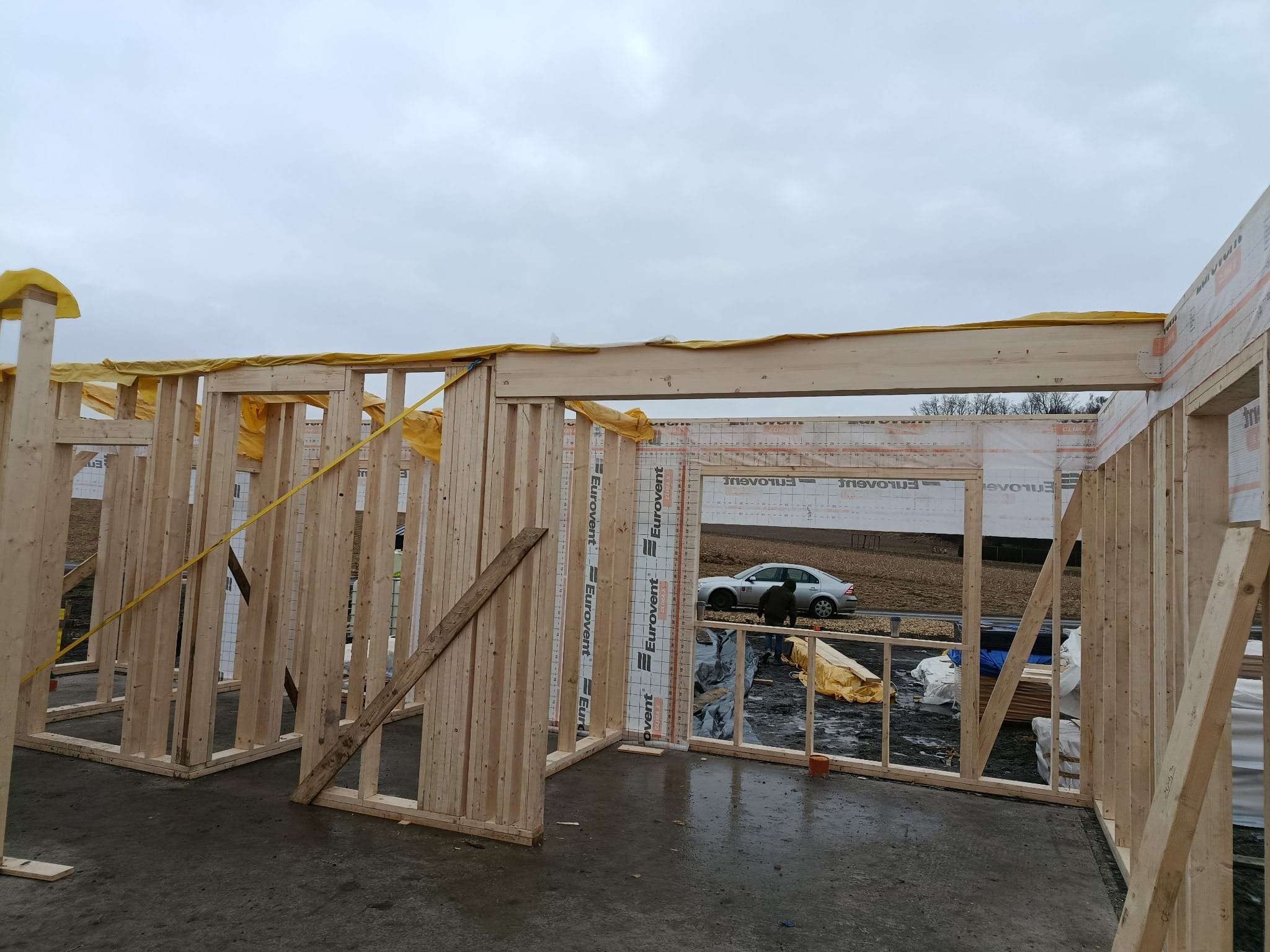 Wnętrze domu szkieletowego w trakcie budowy. Widoczne drewniane ramy ścian, folia izolacyjna, fragment podłogi i samochód w tle. Pochmurne niebo.