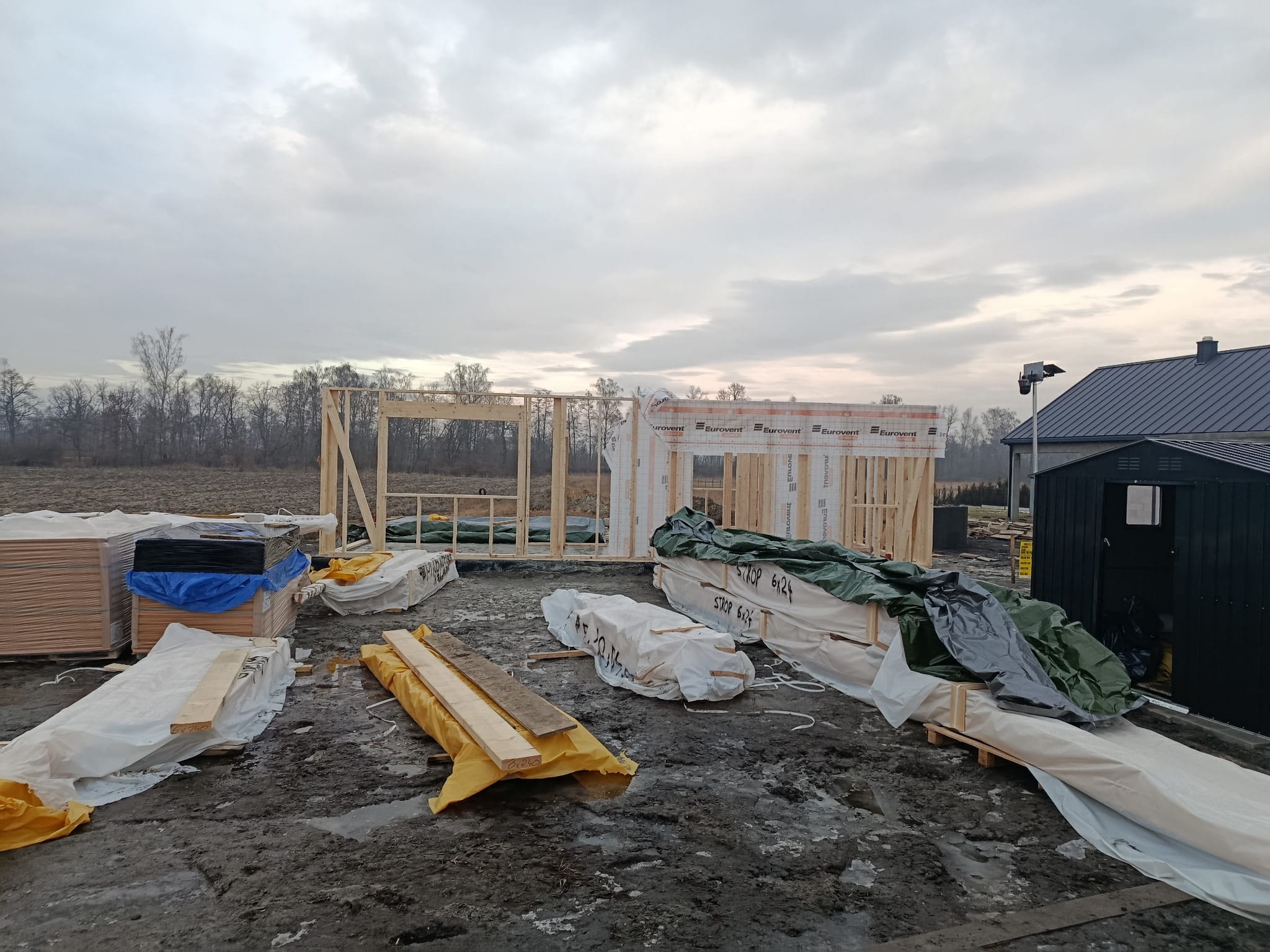Wczesny etap budowy: szkielet domu z drewnianych belek na fundamencie, materiały budowlane zabezpieczone folią, w tle widoczne drzewa i budynek z ciemnym dachem.