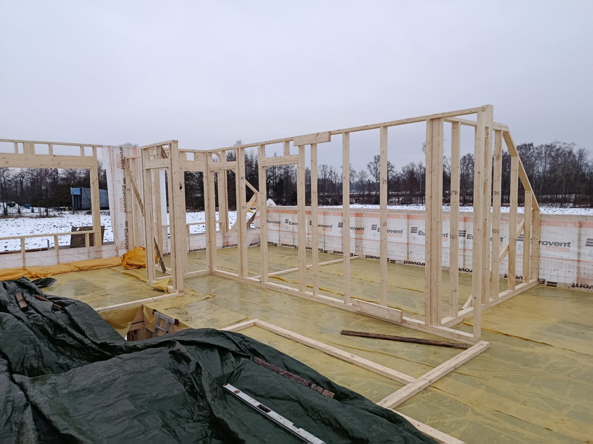 Wnętrze w trakcie budowy: drewniany szkielet domu z widoczną izolacją Eurovent, poziomica na folii ochronnej, zimowy krajobraz w tle. Etap wznoszenia ścian.
