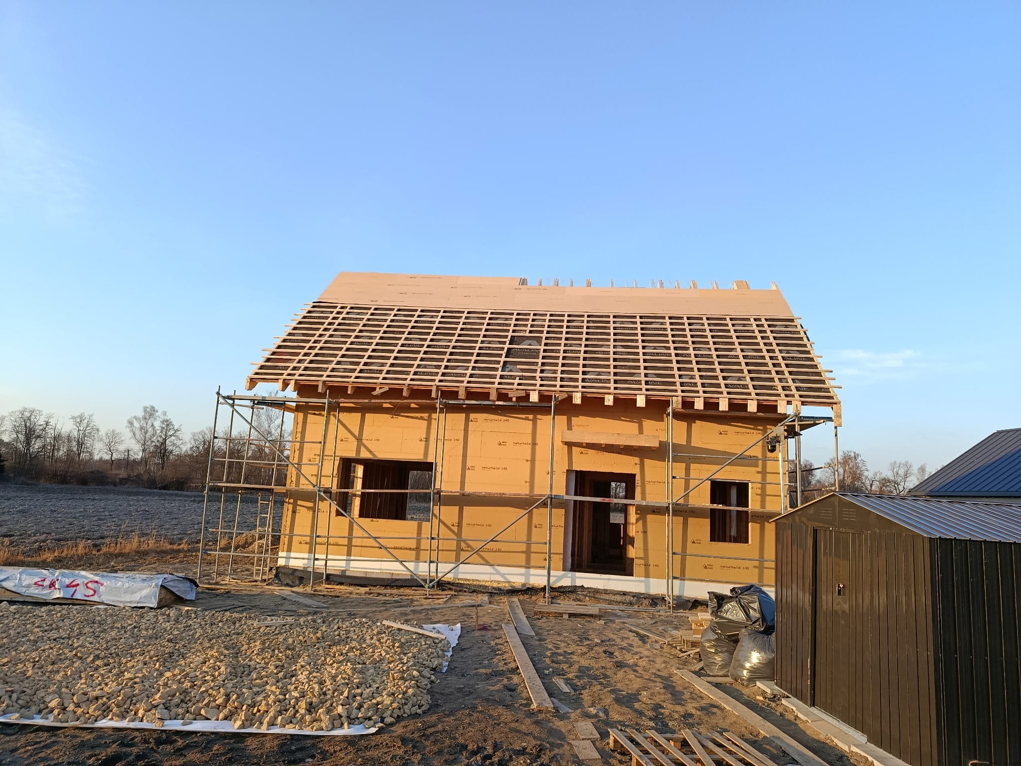 Budowa domu z drewnianą konstrukcją dachu w trakcie realizacji. Widoczne rusztowania, materiały budowlane i elementy konstrukcyjne. Jasne niebo w tle.